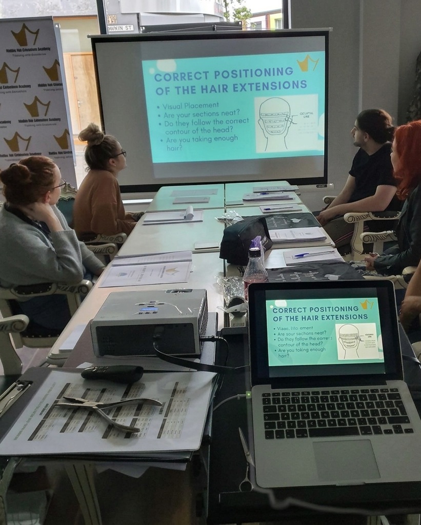 Students seated around a classroom table viewing a projected lesson on hair extension positioning at iHobbs Academy.