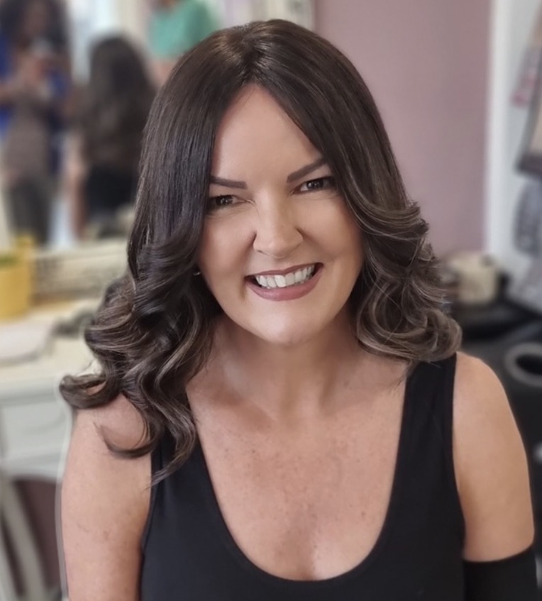 A woman with long, dark wavy hair curled at the ends, smiling in the iHobbs clinic, wearing a finished Custom Wig.