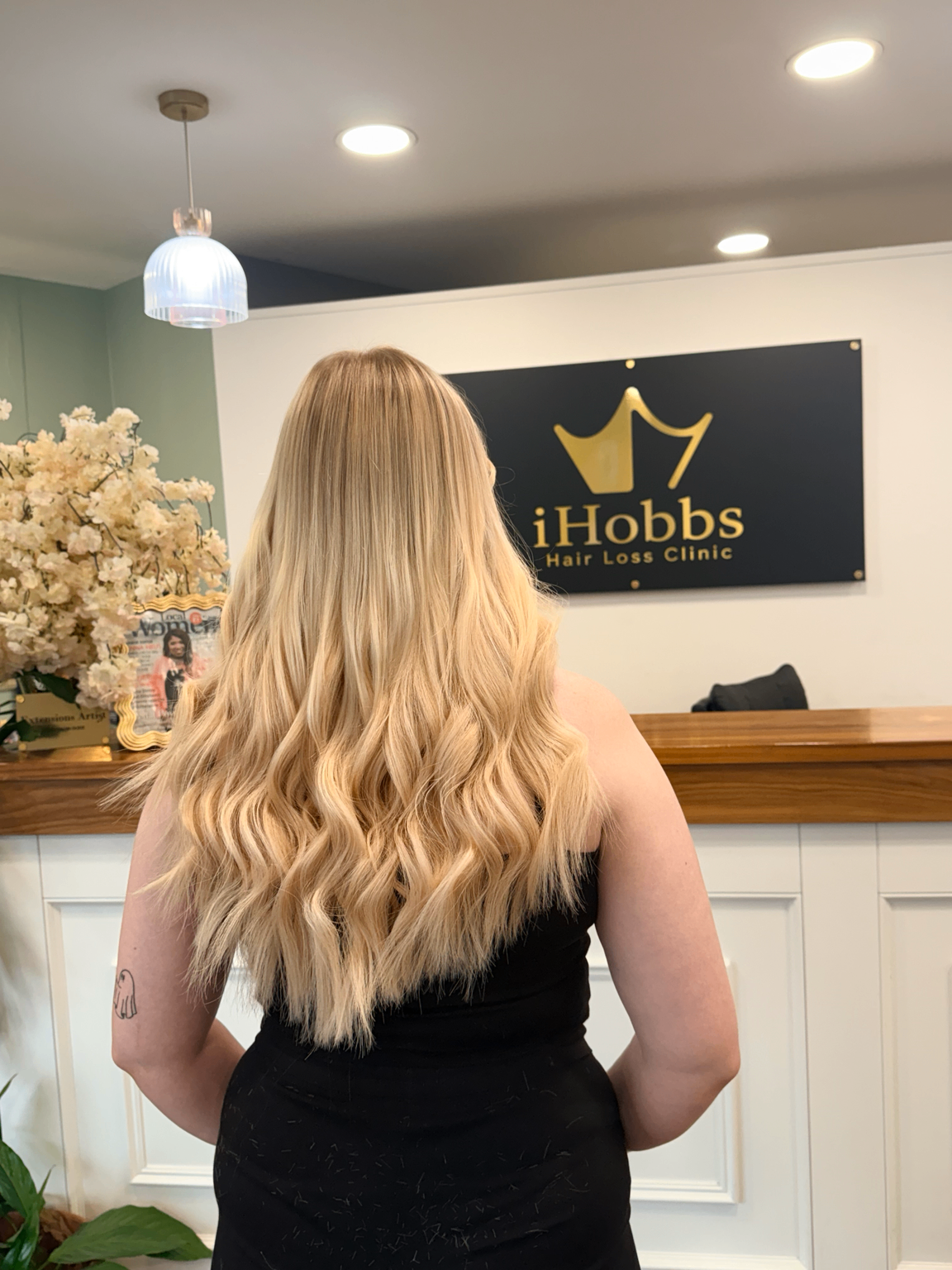 Long blonde hair finished in soft waves, photographed from behind in front of the iHobbs Hair Loss Clinic reception sign.