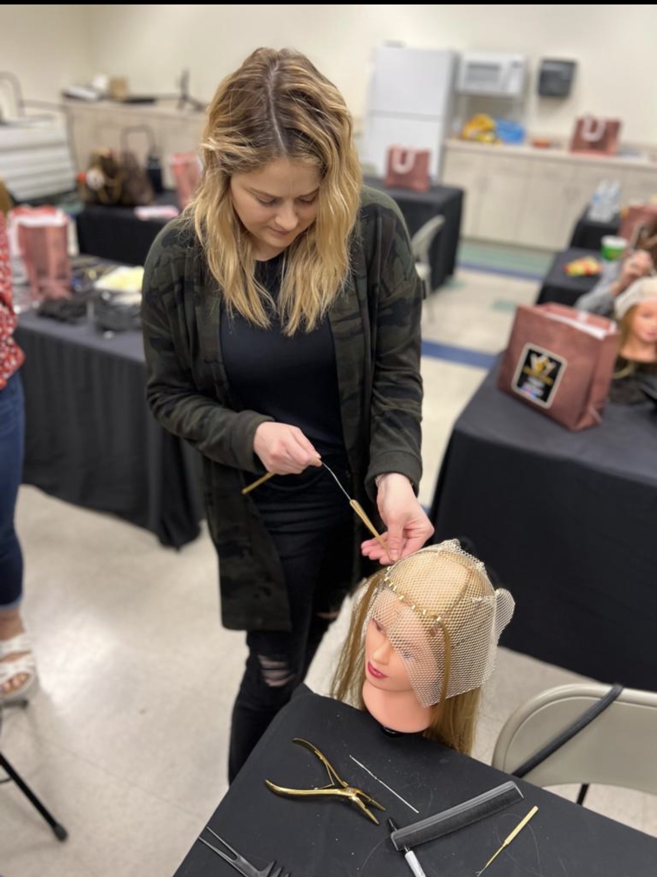A practitioner ventilating a fine mesh hairpiece base onto a mannequin head with a latch hook tool in the iHobbs Academy training room.